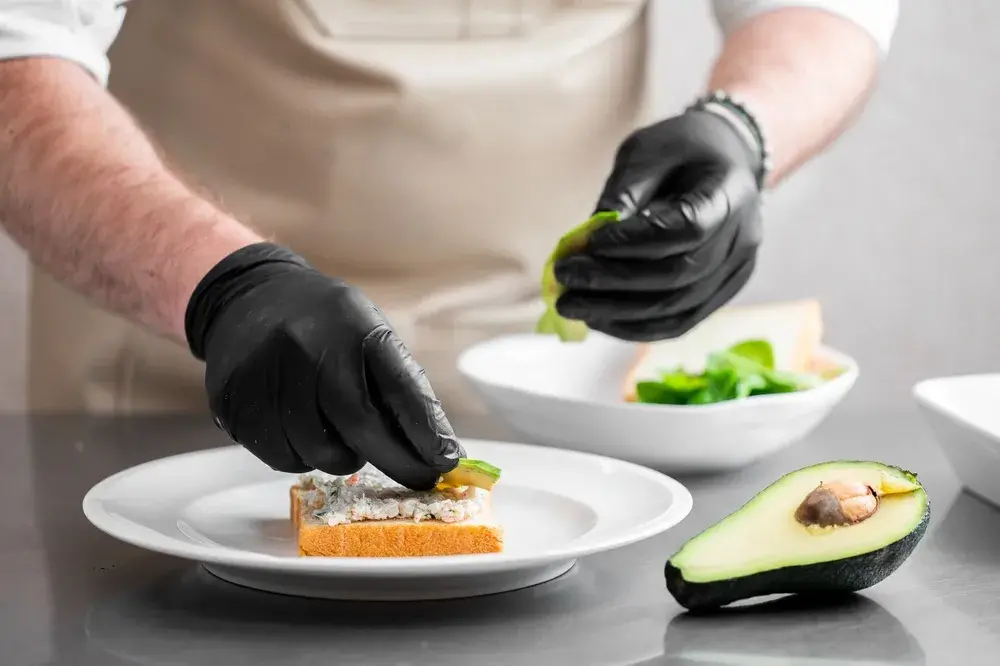Hands wearing black gloves assembling a snack with leafy greens on a spread-covered slice of bread, with half an avocado nearby on a kitchen counter.