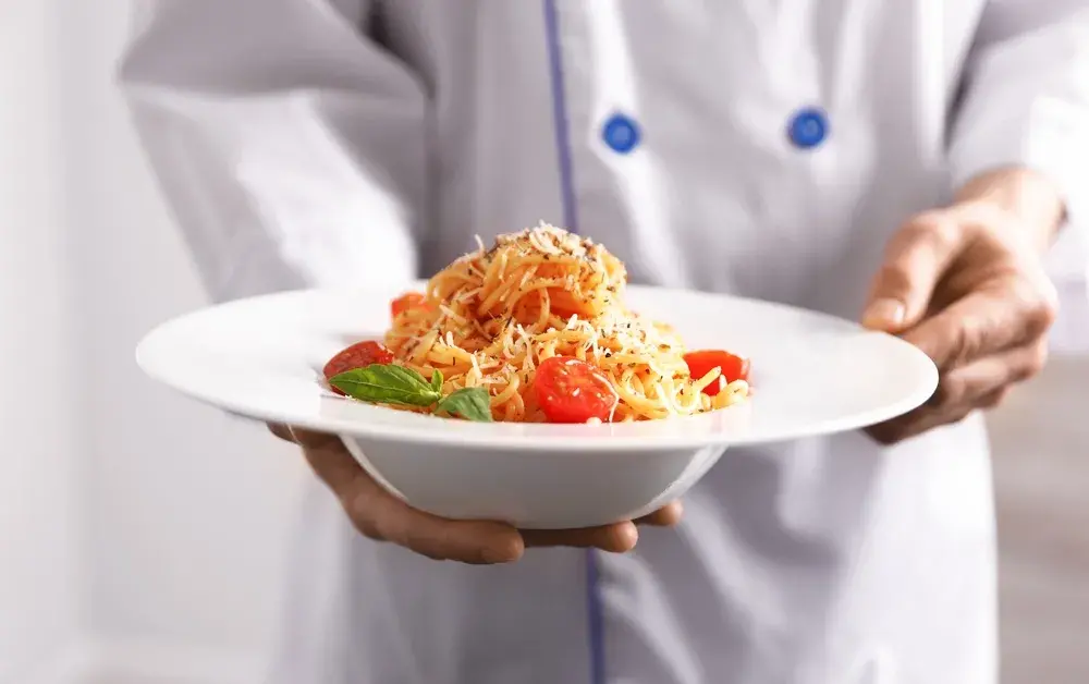 Chef holding a plate of spaghetti pasta garnished with cherry tomatoes, basil, and shredded cheese in a dining setting.